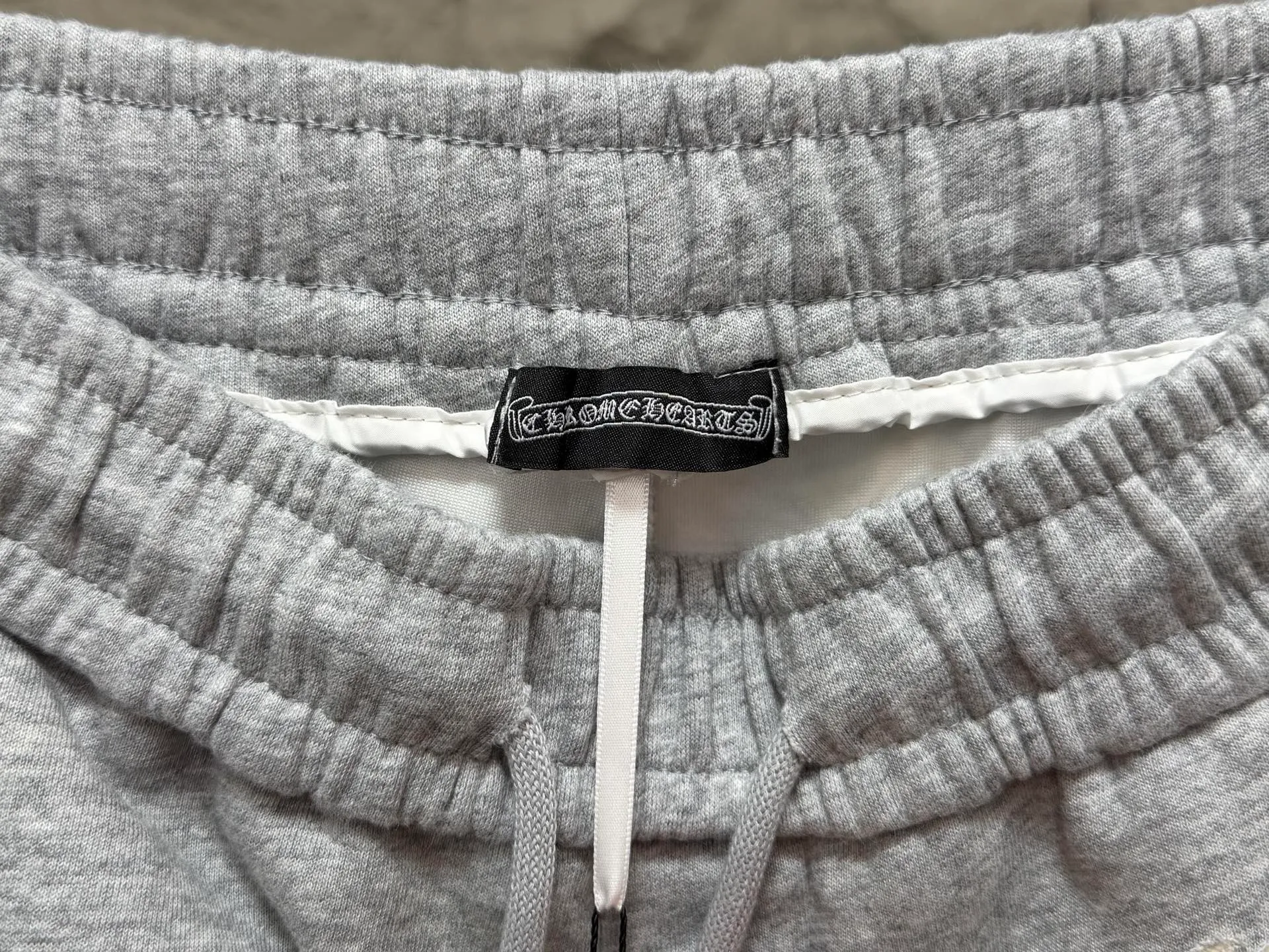 CH/Cro Heart 2026ss Burnt Flower Straight-leg Sweatpants Classic heavy-weight sweatpants with gray tones, Retro street style full-fitting, direct recognition, same style for men and women. ●Three-dimensional patchwork technology: The whole body is covered with Gothic letters + cross flower totems, using raw edge patchwork embroidery, with a mustache-like old texture, and uneven and layered patterns. It is not an ordinary print, and it is durable and not easy to fall off. ●Style tailoring: Loose, straight and wide-legged version, modifying the shape of the legs, hiding the flesh neatly, not flattering the figure, with an online drape, and the upper body has a relaxed street tone. ●Fabric and waist: Heavy knitted terry fabric, thick and soft, with good drape; elastic drawstring waist, with branded metal engraved rope ends, easy to put on and take off, and strong waist adaptability. ●Detailed design: The side pockets are convenient for carrying items, and the back pocket is decorated with cross-pattern patches. The front and rear patterns echo each other. Every detail is highly recognizable to the brand. ●Wearing scenarios: Suitable for street wear, daily outings, and trendy wear. Paired with sweaters, denim jackets, and leather jackets, you can easily create an American retro cool style. ●The overall gray base is paired with Cream White patches, which are low-key yet impactful. They are both comfortable and trendy. Versatile sweatpants, a must-have for fashionistas, are also available at the counter. SIZE：M L XL XXL XXXL